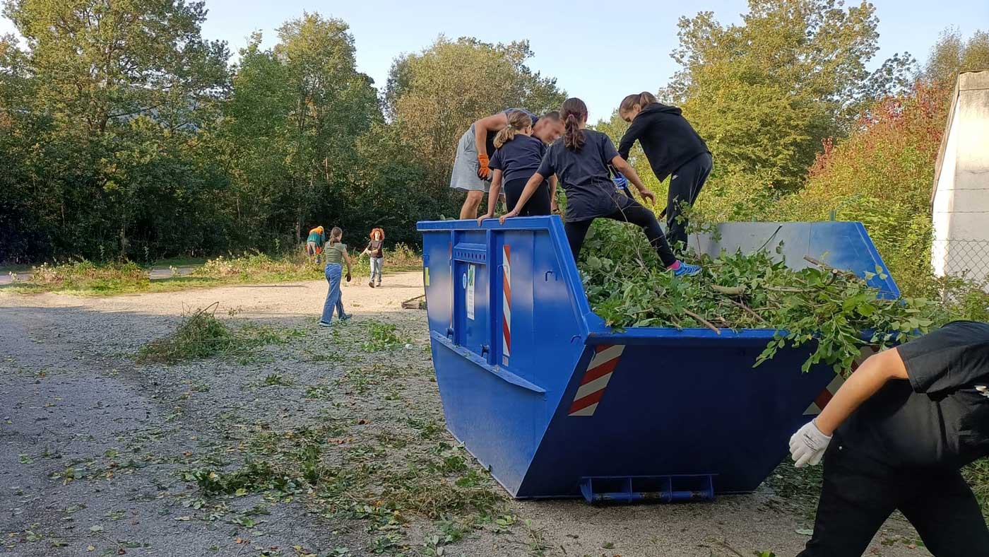 Kinder in Frankfurt füllen eifrig einen Container mit Grünabfall, demonstrierend wie der Containerdienst Frankfurt zur Umwelterziehung beiträgt