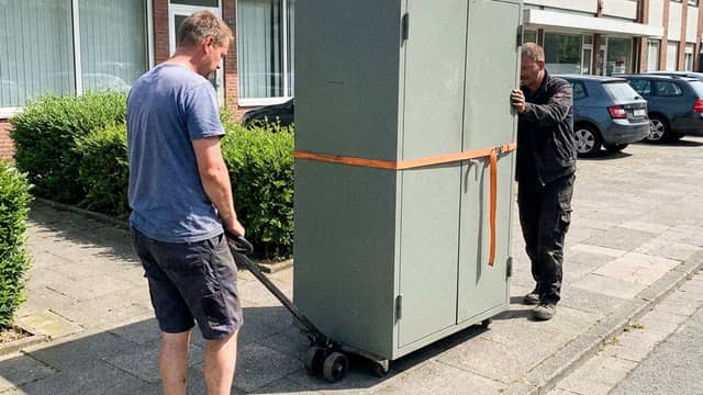 Zwei Mitarbeiter schieben einen gesicherten Doppeltür-Wertschrank auf einem Schwerlastroller über den Gehweg Zwei Mitarbeiter schieben einen gesicherten Doppeltür-Wertschrank auf einem Schwerlastroller über den Gehweg